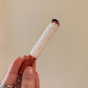 May include: A beaded keychain in the shape of a cigarette. The keychain is white with iridescent beads, an orange-brown base, and a black tip. The keychain is held by a hand.
