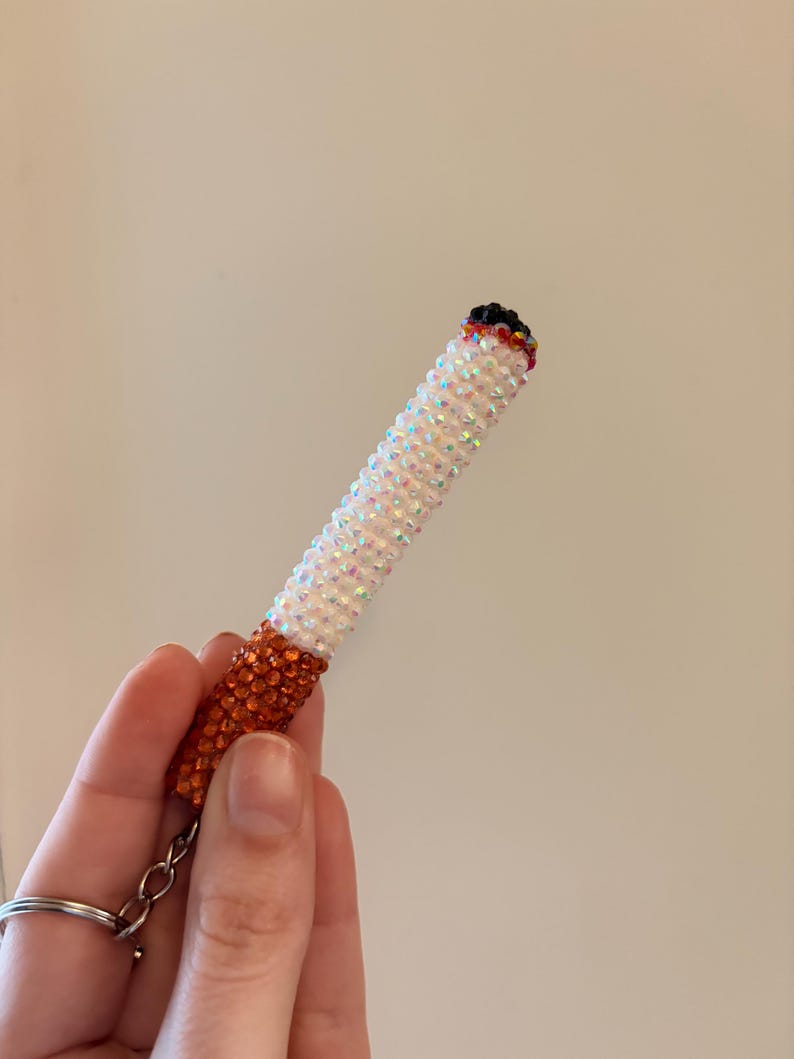May include: A beaded cigarette-shaped keychain. The keychain features white iridescent beads, an orange-brown base, and a black tip. The keychain is held by a hand.