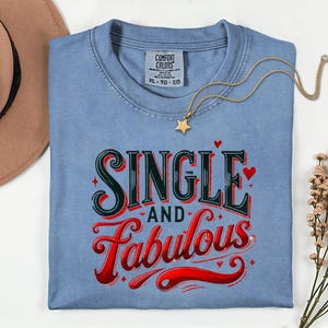 Puede incluir: Camiseta azul claro con la frase "SINGLE and Fabulous" en un diseño de letras rojas y negras. Un collar dorado con un colgante de estrella descansa sobre la camiseta. Un sombrero marrón y flores en el fondo.