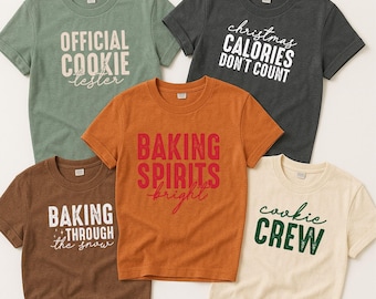 Peut inclure: Cinq t-shirts à manches courtes de différentes couleurs, chacun avec un motif de texte sur le thème des fêtes. Les chemises sont dans les tons de vert, marron, orange, gris et crème. Le texte comprend des phrases comme "Official Cookie Tester" et "Christmas Calories Don't Count."