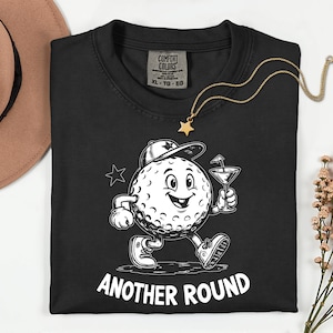 May include: Black t-shirt with a cartoon golf ball character wearing a cap and holding a cocktail. The text "ANOTHER ROUND" is printed in white. A gold star necklace is draped across the shirt.