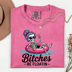 Puede incluir: Una camiseta rosa con el gráfico de un esqueleto con pelo azul y rosa y gafas de sol, relajándose en un flotador rosa mientras sostiene una botella. El texto dice Bitches BE FLOATIN. La camiseta tiene una etiqueta Comfort Colors en talla XL.
