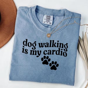Puede incluir: Camiseta azul claro con la frase "dog walking is my cardio" en letras negras, acompañada de dos huellas de patas negras. Un collar dorado con un colgante descansa sobre la camiseta. La camiseta está doblada y un sombrero marrón es parcialmente visible.