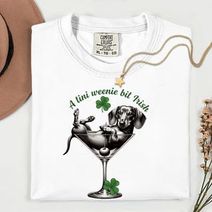 May include: White t-shirt with a graphic of a dachshund dog relaxing in a martini glass with a cherry. The text "A tini weenie bit Irish" and shamrocks are printed in green. A gold star pendant necklace is draped on the shirt.