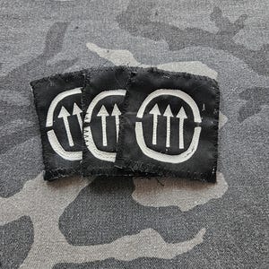Puede incluir: Tres parches cuadrados de tela negra con un diseño blanco de tres flechas que apuntan hacia arriba dentro de un círculo. El fondo es un patrón de camuflaje gris.