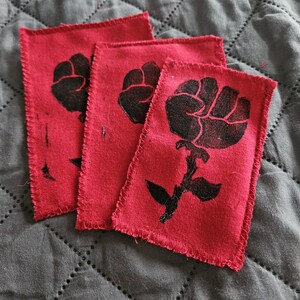 May include: Three red fabric patches with a black fist and rose design. The patches have a raw edge and are arranged on a gray quilted surface. The fist and rose design is a symbol of solidarity.