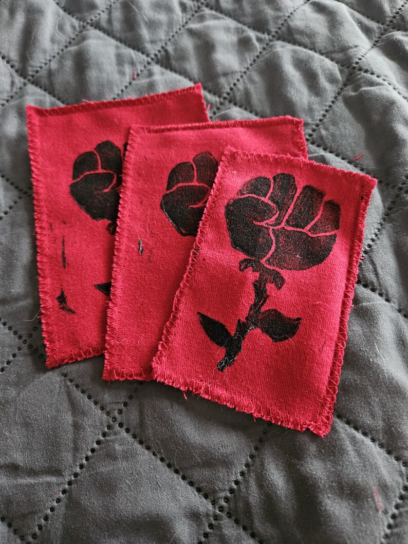 May include: Three red fabric patches with a black fist and rose design. The patches have a raw edge and are arranged on a gray quilted surface. The fist and rose design is a symbol of solidarity.