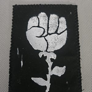 May include: A black square patch with a white fist-shaped flower design. The patch has a stitched border and a textured appearance. The design is a stylized representation of a fist and a rose.