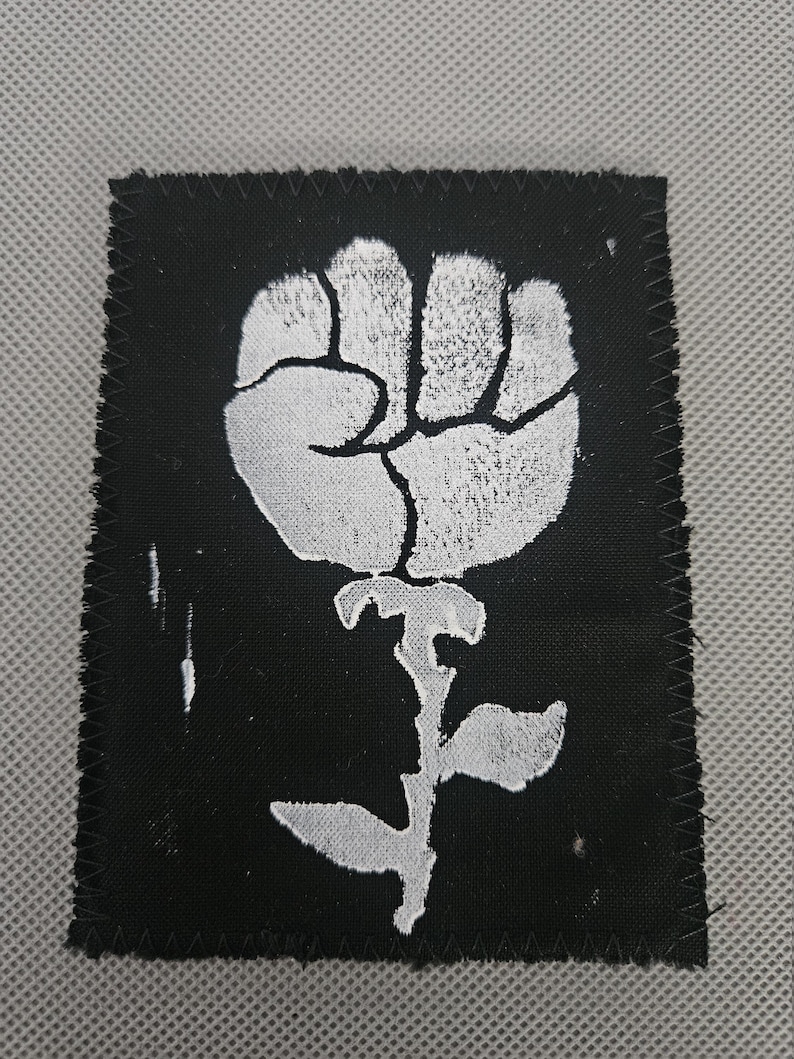 May include: A black square patch with a white fist-shaped flower design. The patch has a stitched border and a textured appearance. The design is a stylized representation of a fist and a rose.