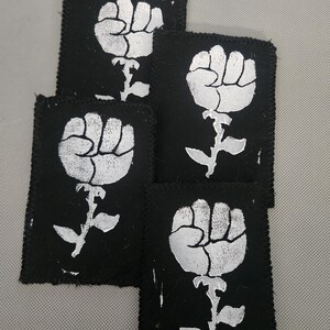 May include: A collection of five black fabric patches. Each patch features a white graphic of a fist emerging from a floral stem. The patches have a stitched border.