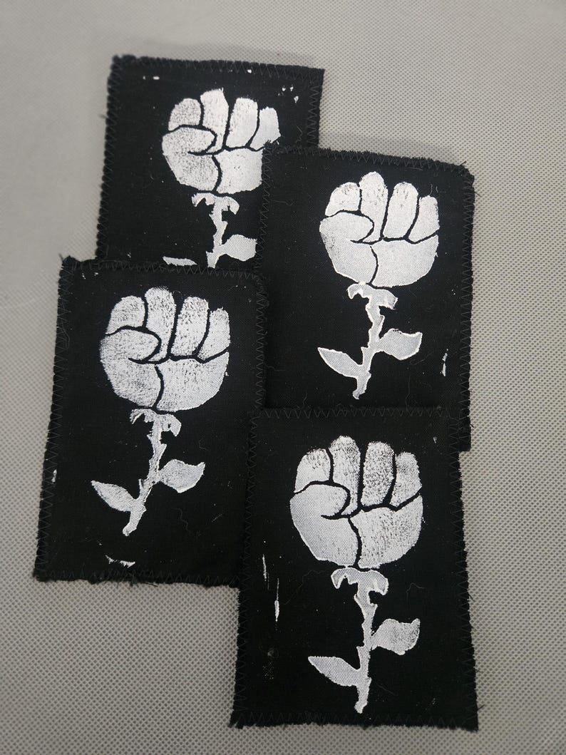 May include: A collection of five black fabric patches. Each patch features a white graphic of a fist emerging from a floral stem. The patches have a stitched border.