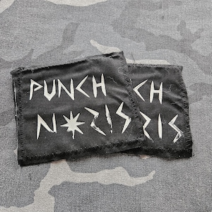 May include: Two black fabric patches with white text that reads "PUNCH NAZIS". The patches are rectangular with a stitched border and are set against a camouflage background.