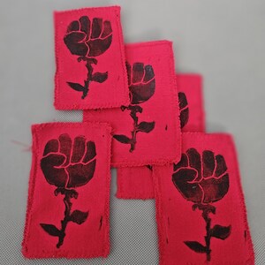 May include: Several red fabric patches with a black fist-and-rose design. The patches have a rectangular shape with frayed edges. The design features a clenched fist atop a stem with leaves, resembling a rose. The patches are arranged in a slightly overlapping pattern.