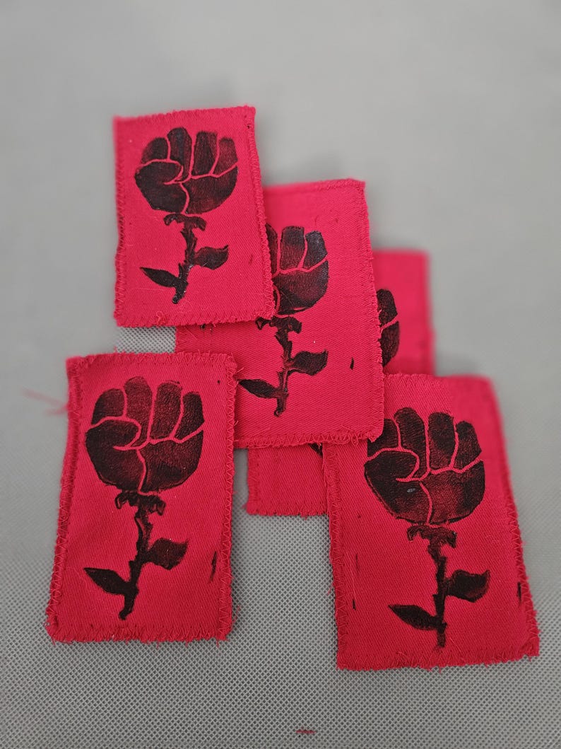 May include: Several red fabric patches with a black fist-and-rose design. The patches have a rectangular shape with frayed edges. The design features a clenched fist atop a stem with leaves, resembling a rose. The patches are arranged in a slightly overlapping pattern.