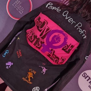 May include: Black denim jacket with a pink patch featuring a raised fist and the words "RESISTS" and "And". The back of the jacket has the words "People Over Profit" and various graphic symbols.