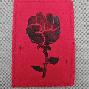 May include: A red fabric patch with a black graphic of a fist rising from a stem with leaves. The patch has a rectangular shape with a stitched border. The design is a symbol of solidarity and resistance.