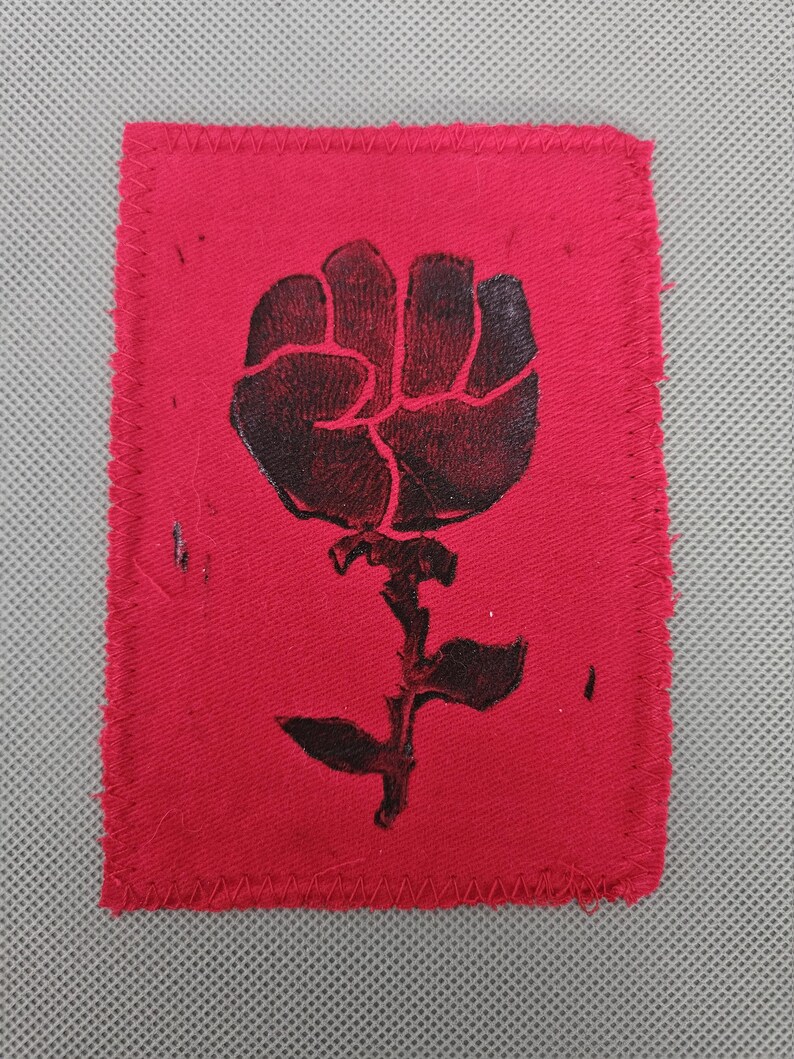 May include: A red fabric patch with a black graphic of a fist rising from a stem with leaves. The patch has a rectangular shape with a stitched border. The design is a symbol of solidarity and resistance.