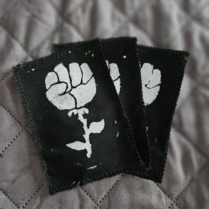 May include: Three black fabric patches with a white fist-shaped flower design. The patches have a stitched border and are arranged in a slightly overlapping pattern. The background is a gray, quilted fabric.