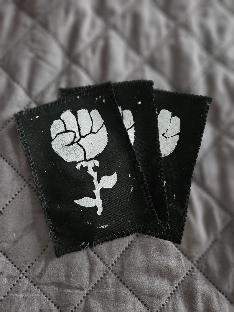 May include: Three black fabric patches with a white fist-shaped flower design. The patches have a stitched border and are arranged in a slightly overlapping pattern. The background is a gray, quilted fabric.