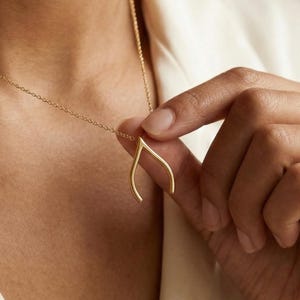 Op de afbeelding: Een delicate goudkleurige ketting met een wensbeen hanger. De hanger hangt aan een fijne ketting. De ketting wordt vastgehouden door een persoon die een crèmekleurige top draagt. De sieraden zijn eenvoudig en elegant.