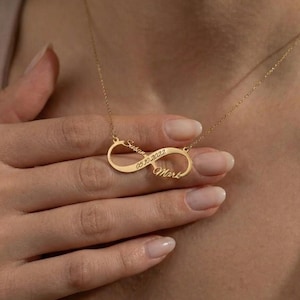 May include: A gold-colored infinity necklace with the names "Susan" and "Mark" engraved on it, along with the date "05.10.2022". The necklace is worn around the neck.