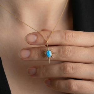 May include: A gold-colored necklace featuring a sunburst pendant with a central oval-shaped blue gemstone. The pendant has radiating spikes and is suspended from a delicate chain. The necklace is displayed against a dark background.