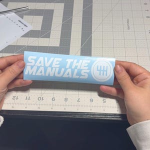 Può includere: Un adesivo azzurro con la scritta bianca "SAVE THE MANUALS" e una grafica bianca di un cambio. L'adesivo è tenuto in mano su un tappetino da taglio grigio.