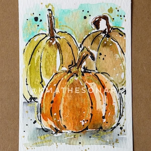 Könnte beinhalten: Aquarellmalerei von drei Kürbissen in Orange-, Gelb- und Brauntönen mit schwarzen Umrissen und goldenen Akzenten. Das Kunstwerk befindet sich auf einem weißen Hintergrund mit einer türkisfarbenen Waschung und dem Text "@KMATHESONART".