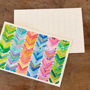 May include: A rectangular greeting card featuring a vibrant watercolor chevron pattern in shades of green, blue, pink, orange, and yellow. The text @kmathesonart is printed in white across the center. A matching envelope with a faint chevron pattern.