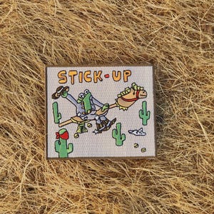 Może przedstawiać: Kwadratowa naszywka z pomarańczowym napisem "STICK-UP". Naszywka przedstawia kreskówkową żabę jadącą na koniu na patyku, z kaktusami i elementami pustynnymi. Żaba ma na sobie kapelusz kowbojski i buty.