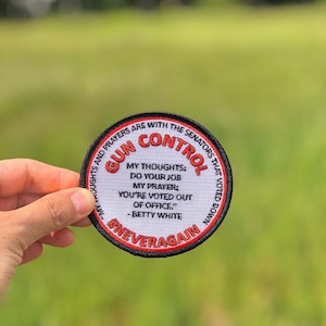 Puede incluir: Un parche redondo blanco con costuras negras y rojas. El texto en el parche dice "Gun Control" en letras rojas con las palabras "My thoughts: Do your job. My prayer: You're voted out of office." - Betty White. El texto "#NeverAgain" está en la parte inferior del parche.