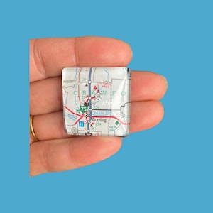 May include: A square, clear glass paperweight featuring a map of Grayling, Michigan. The map includes roads, highways, and place names in a colorful design. The paperweight is held in a hand against a blue background.
