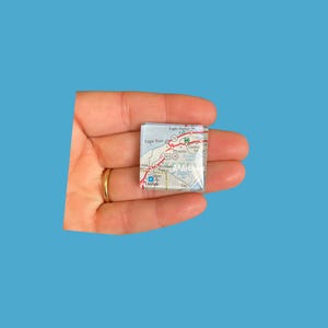 May include: A small, square map tile featuring a detailed road map of a specific area, with place names like Eagle Harbor and Eagle River visible. The tile is held in a hand against a blue background. The map has red and white lines.