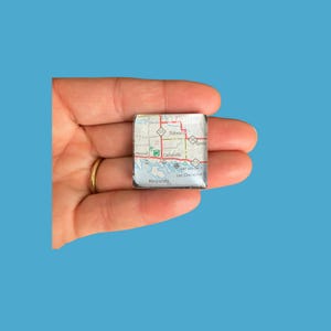 May include: A square, clear glass magnet featuring a map of an area with the names Hessel, Cedarville, and Les Cheneaux. The map is in shades of white, green, and red. The magnet is held in a hand against a blue background.