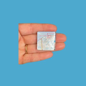 May include: A square, clear glass tile featuring a map design with the text "Lake of the Clouds" and "Porcupine Mths". The map includes red and white lines, and the text "A2 Artworks".
