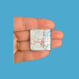 May include: A small, square map image, held in a hand, featuring a detailed map with place names like Keweenaw, Assinins, and L'Anse. The map is predominantly white, green, and red, with a glossy finish. The background is a solid blue.