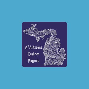 May include: A custom magnet featuring a white outline of the state of Michigan on a dark blue background. The state outline is filled with white text of cities and landmarks. The text "A²Artisans Custom Magnet" is also present.