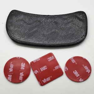 May include: A black, curved, textured plastic component positioned above three adhesive pads. Two pads are circular and one is square, all featuring red backing paper with white 3M VHB text printed repeatedly across the surface.