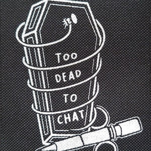 May include: Black square patch with a white coffin design wrapped in a spiral, with the words "TOO DEAD TO CHAT". A white pen and ring are below the coffin. The patch is made of a textured fabric.