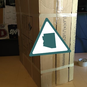 K&ouml;nnte beinhalten: Ein gro&szlig;er, rechteckiger Karton, mit Klebeband befestigt, steht auf einer Holzoberfl&auml;che. Die Box hat aufgedruckten Text und Abmessungen: 52 cm (B) x 23 cm (L) und 62 cm (L) x 52 cm (B) x 8 cm. Ein dreieckiger Aufkleber mit Umriss von Arizona ist sichtbar.