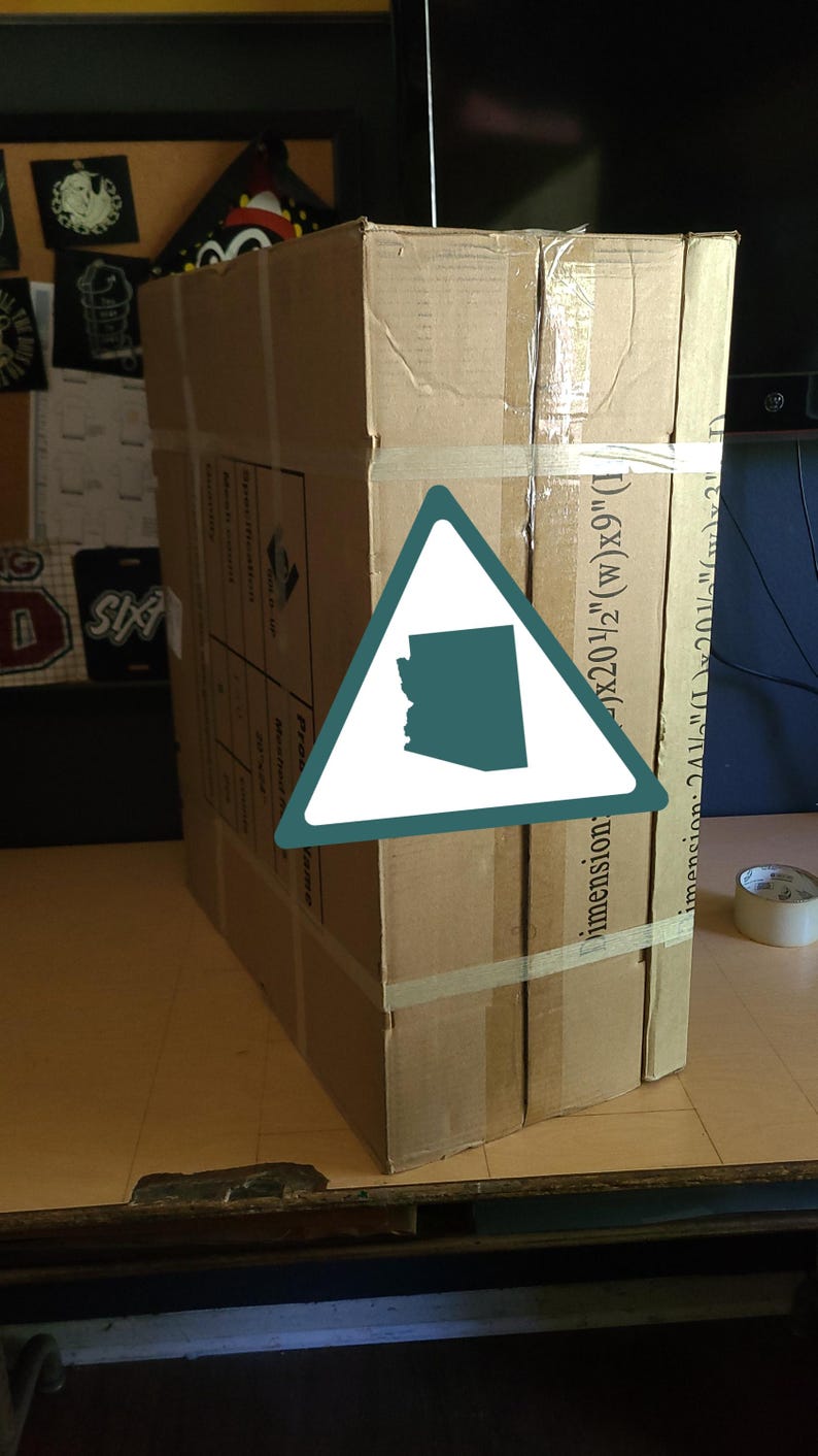 K&ouml;nnte beinhalten: Ein gro&szlig;er, rechteckiger Karton, mit Klebeband befestigt, steht auf einer Holzoberfl&auml;che. Die Box hat aufgedruckten Text und Abmessungen: 52 cm (B) x 23 cm (L) und 62 cm (L) x 52 cm (B) x 8 cm. Ein dreieckiger Aufkleber mit Umriss von Arizona ist sichtbar.