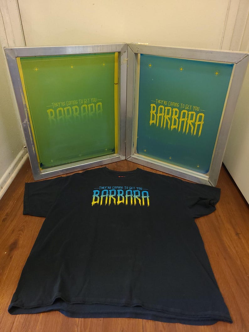 K&ouml;nnte beinhalten: Schwarzes T-Shirt mit dem Text "THEY'RE COMING TO GET YOU BARBARA" in Blau und Gelb. Zwei Siebdruckrahmen befinden sich hinter dem Shirt, einer mit gr&uuml;ner und der andere mit blauer Tinte.