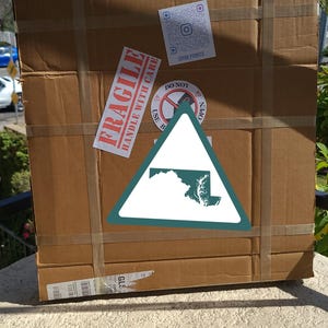 K&ouml;nnte beinhalten: Ein gro&szlig;er, rechteckiger Karton, der mit braunem Klebeband verschlossen ist. Die Box hat mehrere Aufkleber, darunter ein "FRAGILE"-Etikett und ein dreieckiges Schild mit einer Umrisszeichnung des Bundesstaates Maryland. Die Box steht drau&szlig;en auf einer Steinoberfl&auml;che.