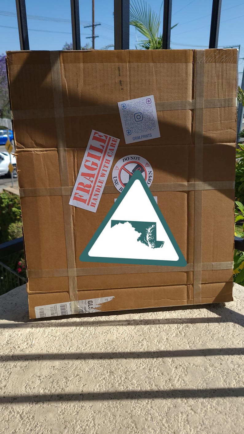 K&ouml;nnte beinhalten: Ein gro&szlig;er, rechteckiger Karton, der mit braunem Klebeband verschlossen ist. Die Box hat mehrere Aufkleber, darunter ein "FRAGILE"-Etikett und ein dreieckiges Schild mit einer Umrisszeichnung des Bundesstaates Maryland. Die Box steht drau&szlig;en auf einer Steinoberfl&auml;che.