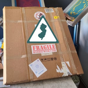 K&ouml;nnte beinhalten: Ein gro&szlig;er, versiegelter Karton mit Klebeband und einem "Zerbrechlich"-Aufkleber. Eine gr&uuml;ne und wei&szlig;e Umrisslinie des Bundesstaates New Jersey befindet sich auf der Box. Ein QR-Code und andere Versandetiketten sind ebenfalls sichtbar.