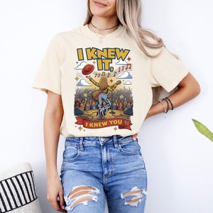May include: A cream-colored short-sleeve t-shirt featuring a graphic of a musician on stage with a guitar, a floating red hat, and musical notes. The text reads I KNEW IT, and I KNEW YOU TS in yellow and red lettering against a concert crowd background.