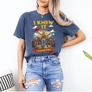 May include: A blue short-sleeve t-shirt featuring a graphic of a performer on stage with a guitar, a red hat, and musical notes. The text reads I KNEW IT, I KNEW YOU TS. The design includes a crowd, stars, and clouds in yellow, red, and white.