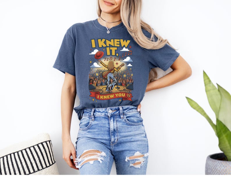 May include: A blue short-sleeve t-shirt featuring a graphic of a performer on stage with a guitar, a red hat, and musical notes. The text reads I KNEW IT, I KNEW YOU TS. The design includes a crowd, stars, and clouds in yellow, red, and white.