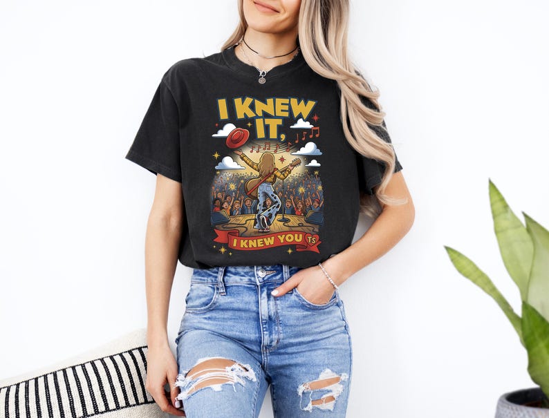 May include: A black short-sleeve t-shirt featuring a graphic of a musician on stage with a guitar, a red hat, and clouds. The text reads I KNEW IT, and I KNEW YOU TS. The design includes musical notes, stars, and a crowd of people in the background.