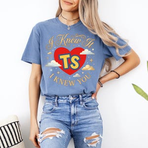 May include: A blue short-sleeve t-shirt featuring a red heart graphic with the yellow text TS inside. The design includes gold cursive text reading I Knew It and I Knew You, surrounded by small white clouds and gold stars printed on the front.